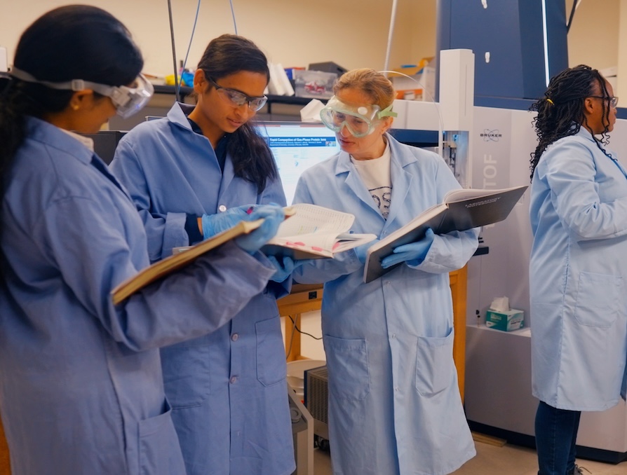 Students in a lab, wearing protective gear, looking at notebooks.