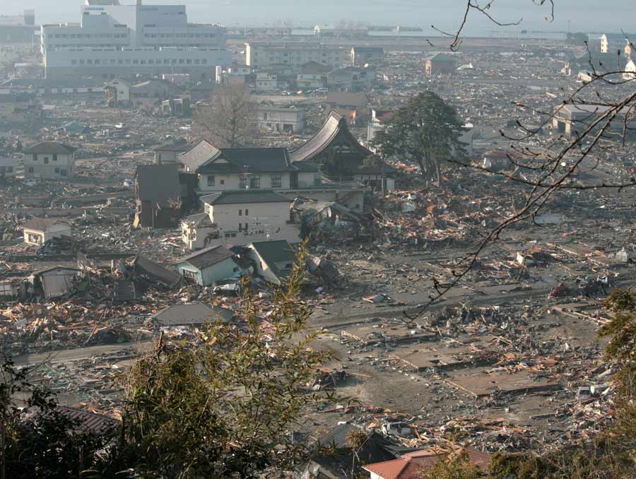 A decimated town after a tsunami with many buildings flattened and destroyed. The ocean is visible in the background.
