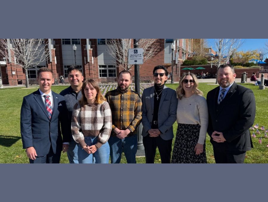 A group of students pose together outside on a sunny day in front of the KC.