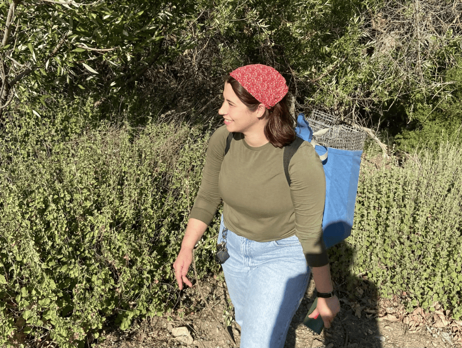 Anita smiles and walks in the sunshine through some bushes with a backpack that has as metal cage in it.