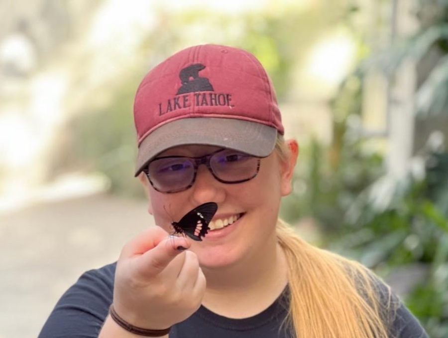 Audrey in a Lake Tahoe hat, smiling at a butterfly that has landed on her finger.