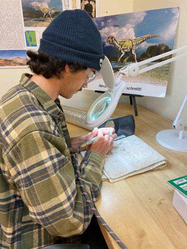 A student wearing safety glasses sits at a desk with a drill working on cleaning a fossil.