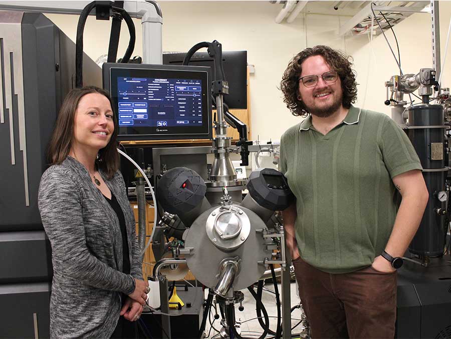 Two people stand next to lab equipment used to research metallic glass.