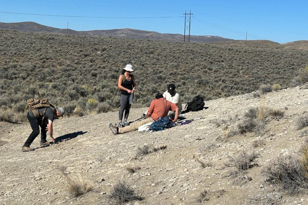 Four people on a bald hillside, one person is standing near two others sitting on the ground looking at the ground between them. Another person nearby is leaning over to pick something up.