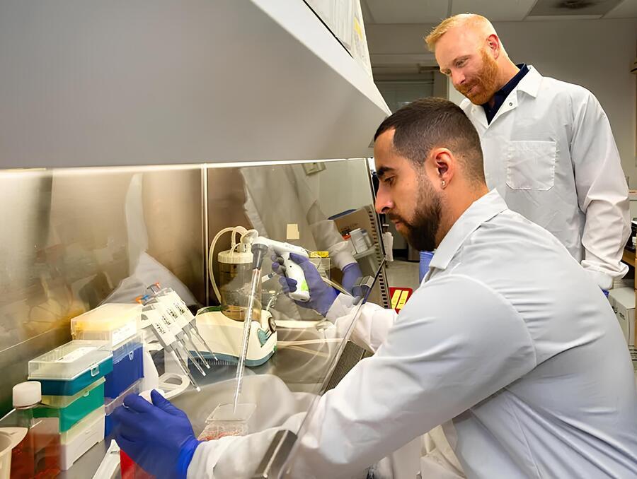 A seated man wearing a white lab coat and blue gloves uses a pipette over a small dish, while another man in a white lab coat stands nearby, observing the process.
