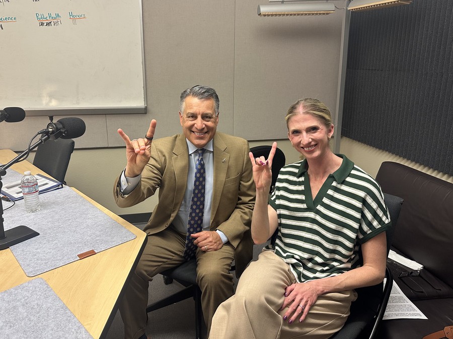 Brian Sandoval sitting next to Jenni Pearson in the podcasting studio holding up Wolf Pack hand signs.