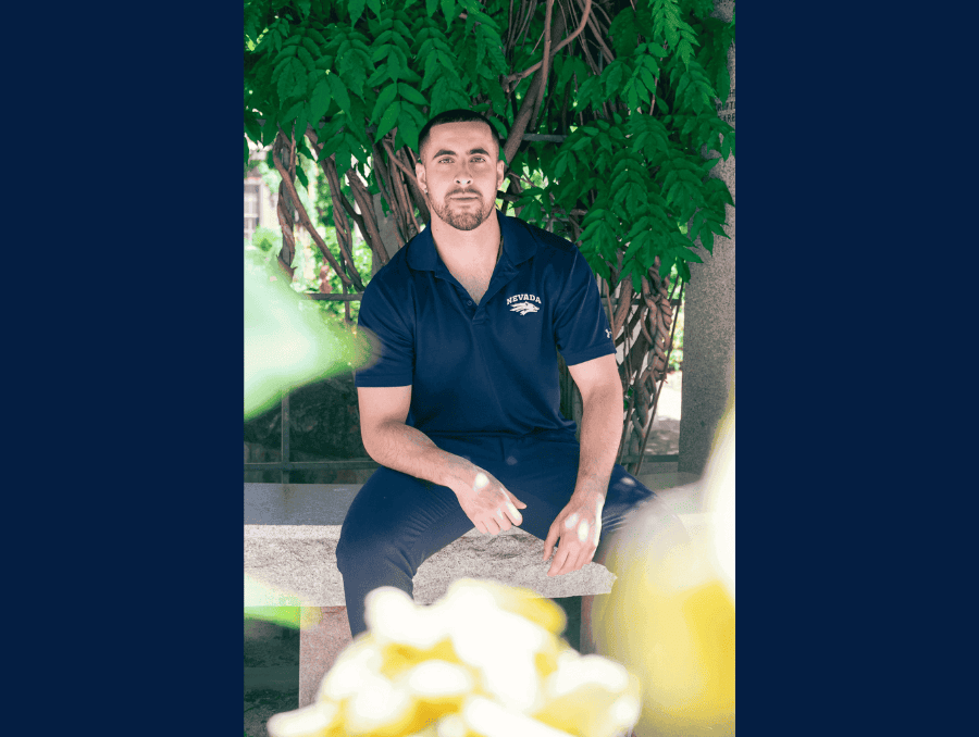 A man in a navy blue T-shirt and pants poses for a photo while seated on a stone bench in front of a leafy tree.