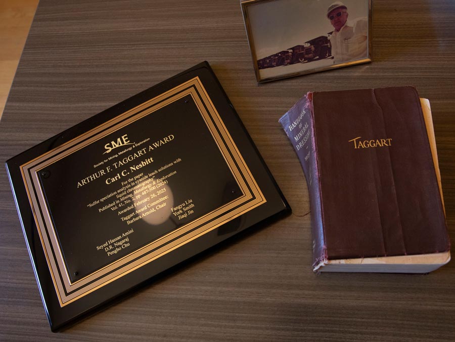 A framed photograph, engraved plaque and tattered book sit on a desk.