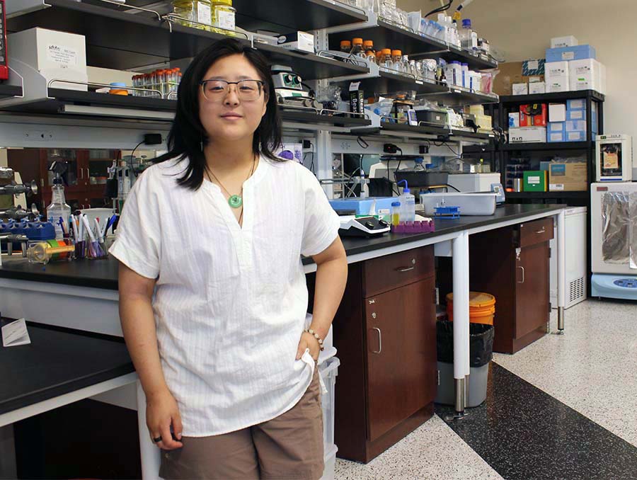 A woman in a white shirt and brown pants stands in front of a lab table with shelves. 