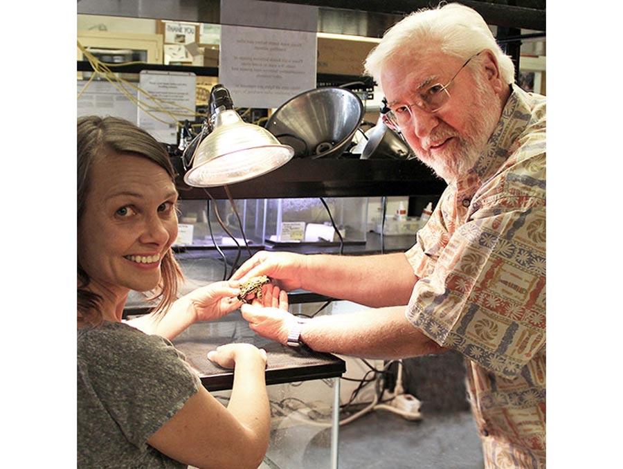 A man, standing, and a woman, sitting, hold a toad and look over their shoulders to smile at the camera.