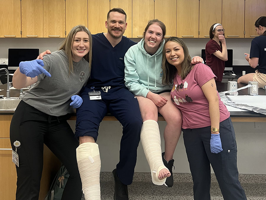 Adam Serfoss and peers learning in the classroom. All are smiling and two of the people have a cast on their leg while sitting on a table.