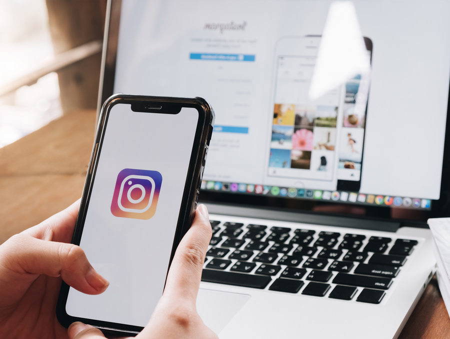 Person holding a smartphone displaying the Instagram logo, with a laptop in the background open to the Instagram login page.