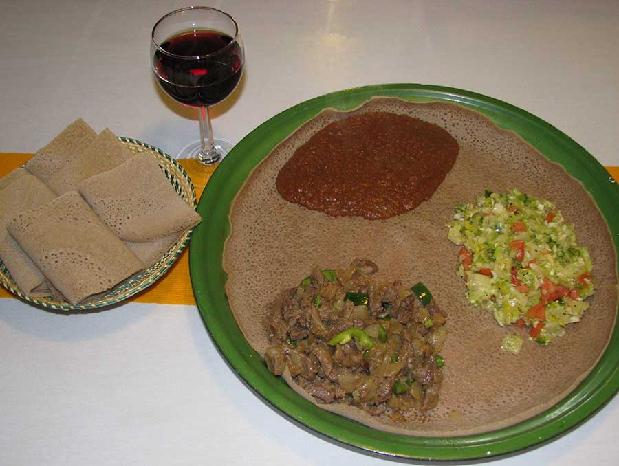 A large, flat injera bread topped with heaping scoops of meat, vegetables and stew is served alongside rolls of injera, as well as a glass of red wine.