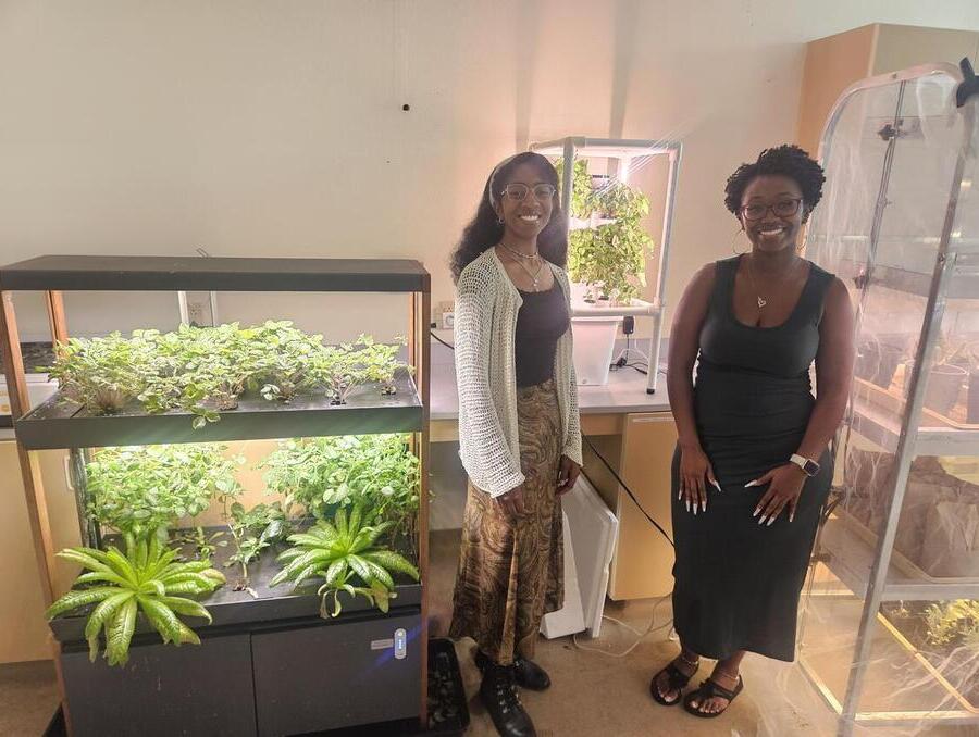 Two smiling women stand beside a glass container filled with growing plants.