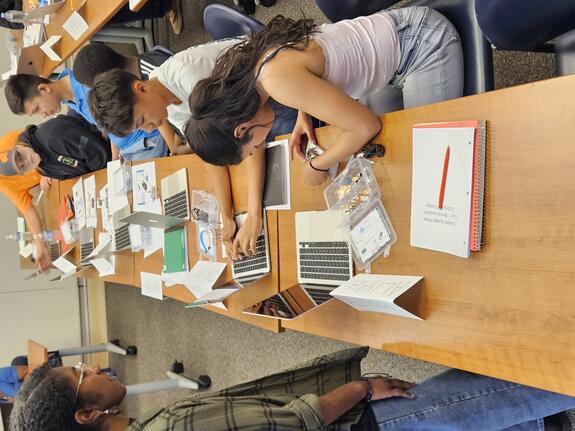 A woman observes a group of boys and girls working on desktop computers, guiding them as they focus on their tasks.