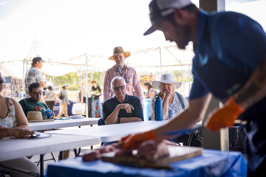 A man wearing a blue apron, orange gloves and holding a knife hunches over a table covered with chunks of meat, demonstrating meat cutting. A small group of men and women sit in front of him, watching closely.