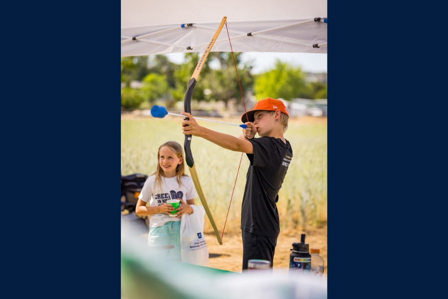 A boy in an orange T-shirt draws a bow, aiming an arrow with focus, while a smiling girl stands nearby, watching in the direction of his aim.