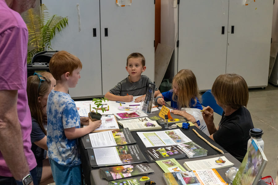 A group of children gathers around a table covered with papers featuring colorful pictures. One child stands holding a small potted plant, while the others sit and write with crayons.