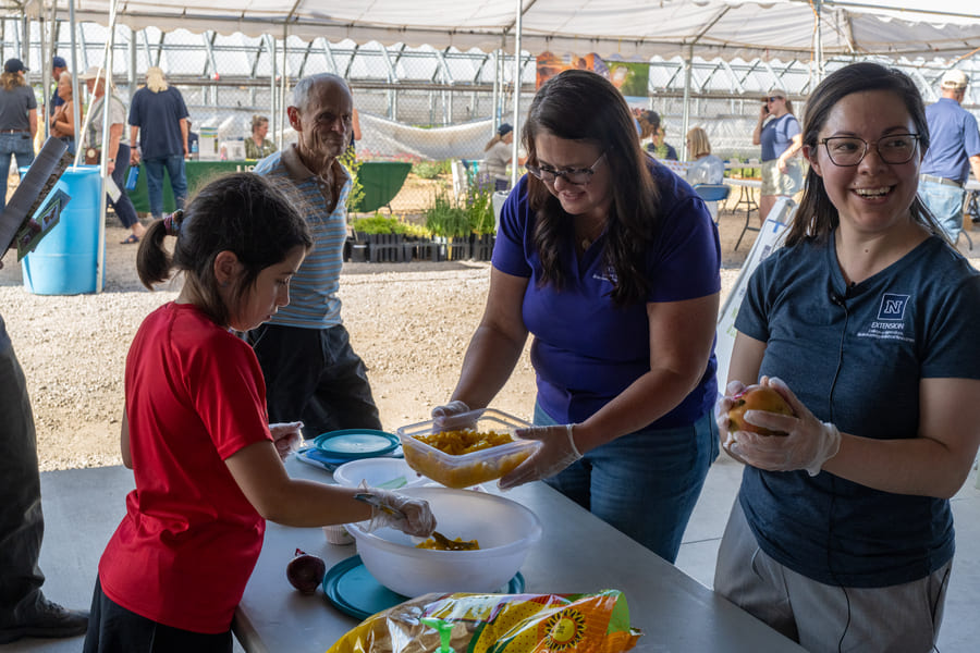 A girl in a red T-shirt and gloves lifts a spoonful of food from a large bowl on a table, while a woman next to her holds a storage container filled with food. Another woman stands nearby, smiling as she looks in a different direction, while a man watches her with a smile.