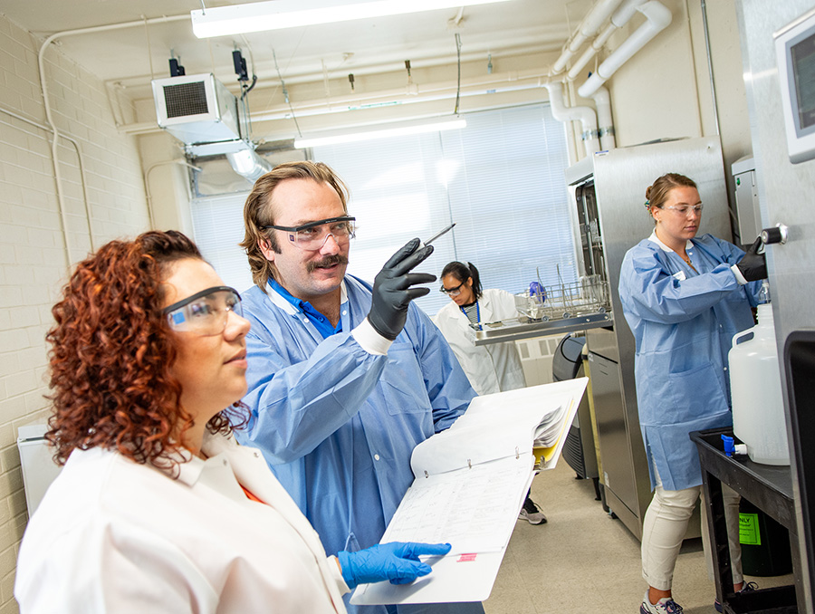 Four laboratory technicians in lab coats are attentively working with scientific equipment in a laboratory setting. One is analyzing a sample in a test tube, two are recording data, and another is operating a machine.