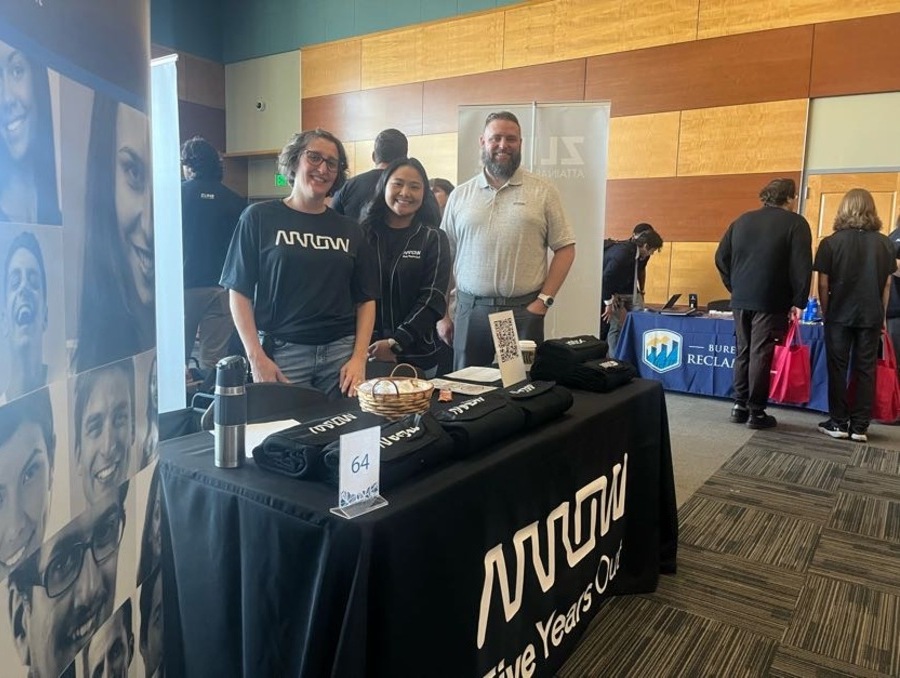 Three Arrow Electronics representatives stand behind a booth at a university career fair, smiling and engaging with attendees. The table displays company-branded materials and a sign reading “Five Years Out.”