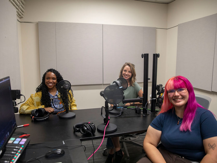Three women sit in a podcasting studio smiling for the camera.