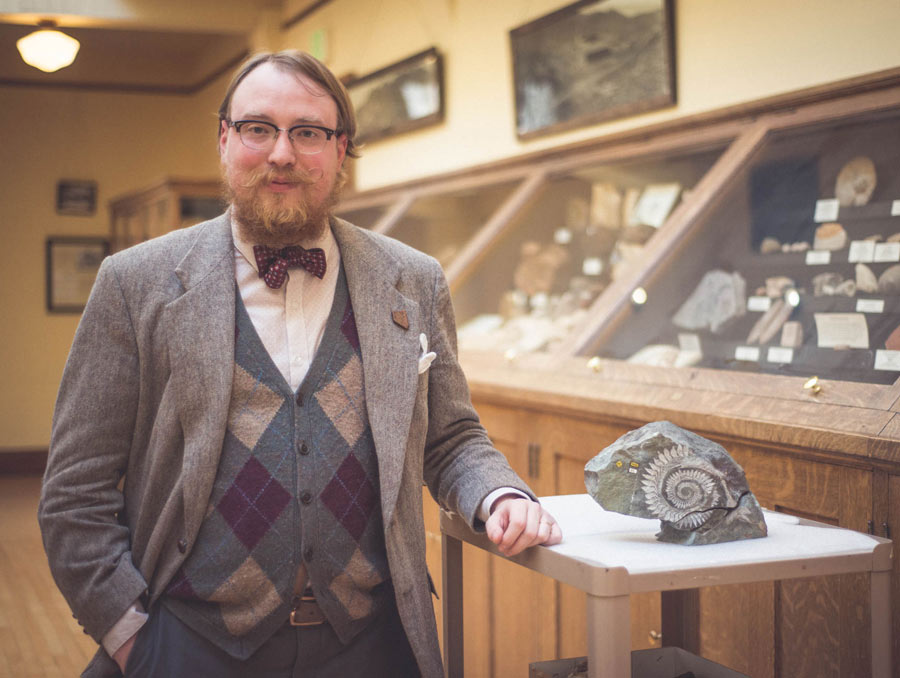 Garrett Barmore stands in the Keck Museum next to a fossil specimen. Barmore is dressed in a bow tie, sweater vest and wool coat with a Nevada lapel pin.
