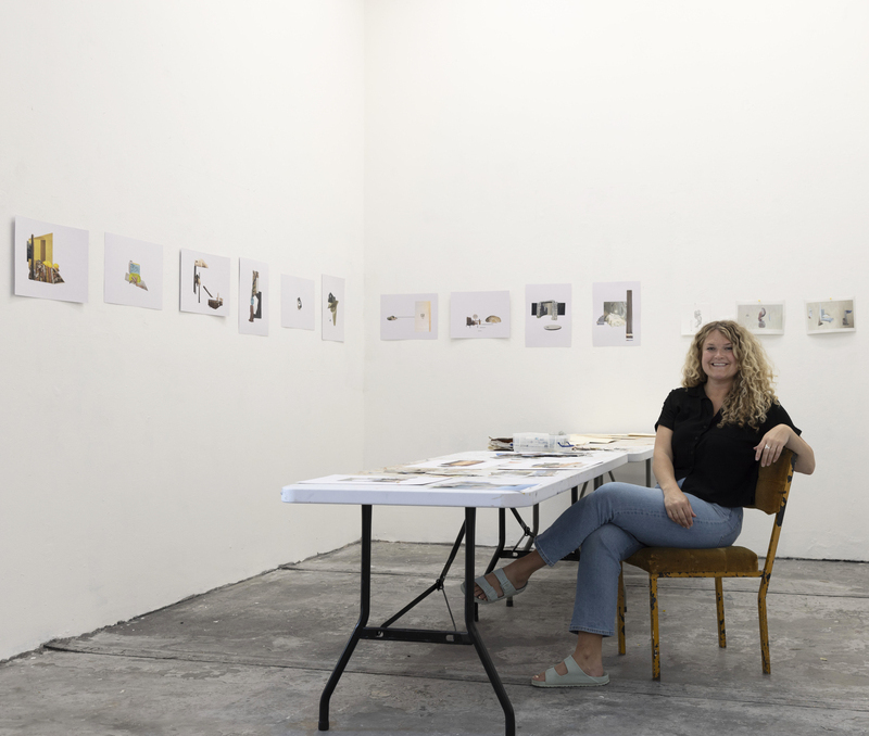 A woman sits smiling at a table with her artwork on the walls surrounding her.