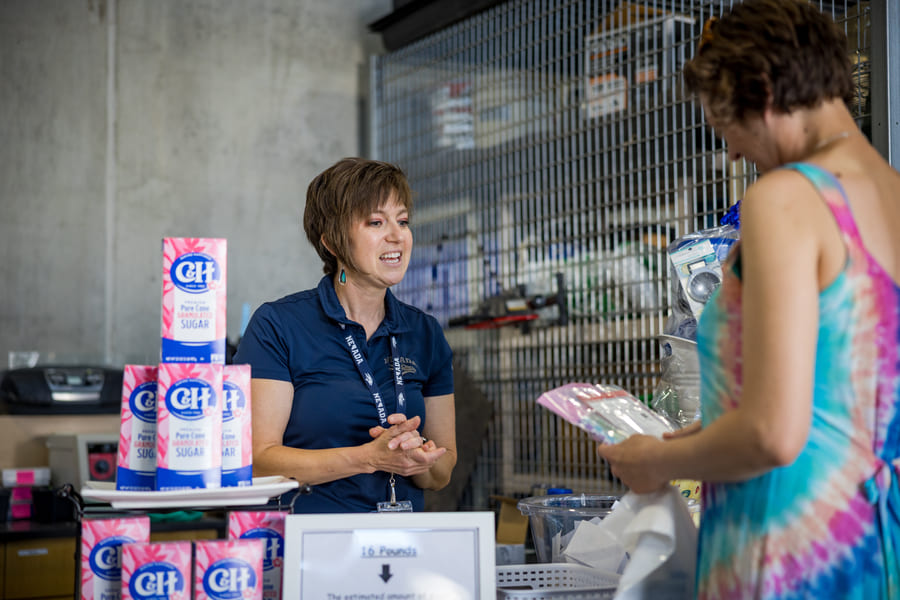 A woman stands behind a table stacked with pink, blue and white boxes of sugar, engaged in conversation with another woman across from her who holds a plastic bag filled with cards.