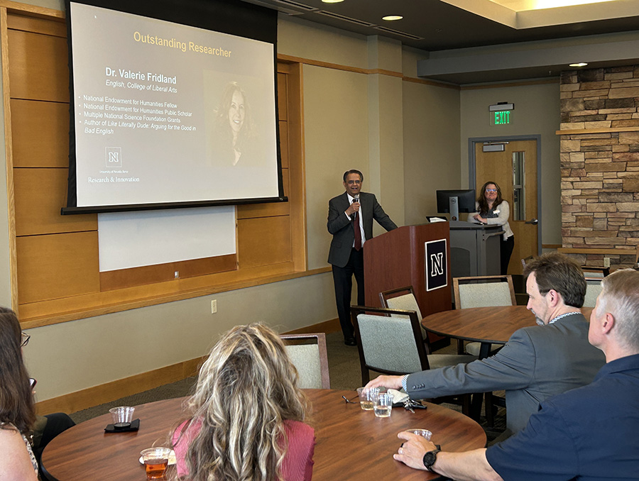 An awards ceremony at the University of Nevada, Reno displaying a presentation slide about Dr. Valerie Fridland, who is recognized as an Outstanding Researcher. Mridul Gautam is speaking at the podium with a presentation slide displayed. The audience listens attentively.