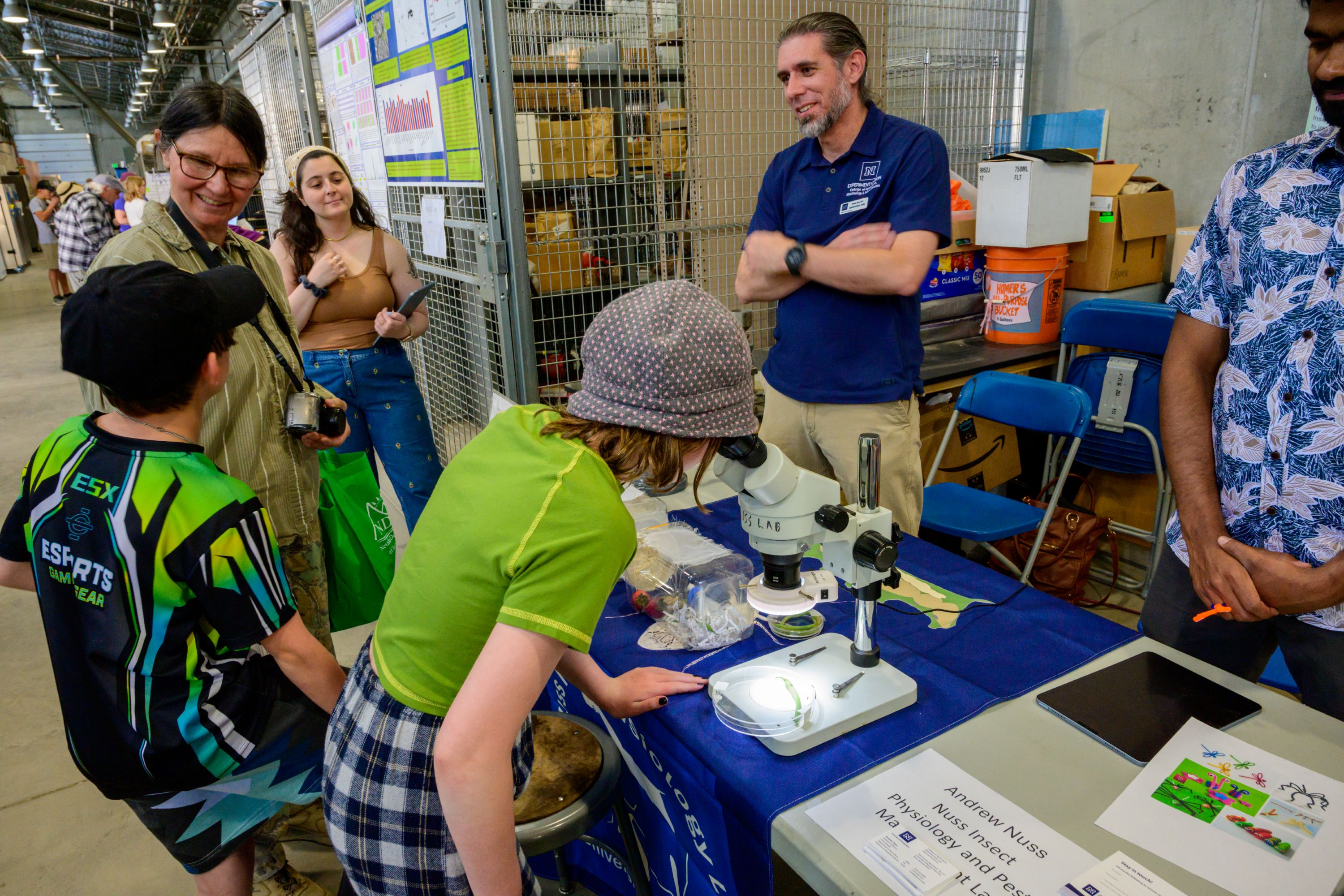Men, women and children standing around a table with one girl looking at a dish through a microscopic lens.