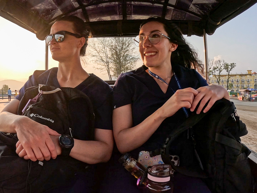 Two nursing students riding in the back of a tuk-tuk in Kampot, Cambodia while holding backpacks on their laps.