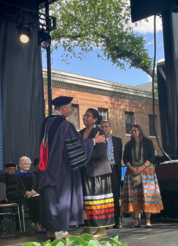 Sandoval stands on the commencement stage with two members of the Burns family as they look at one another and accept the honorary doctorate on Ralph Burns' behalf.