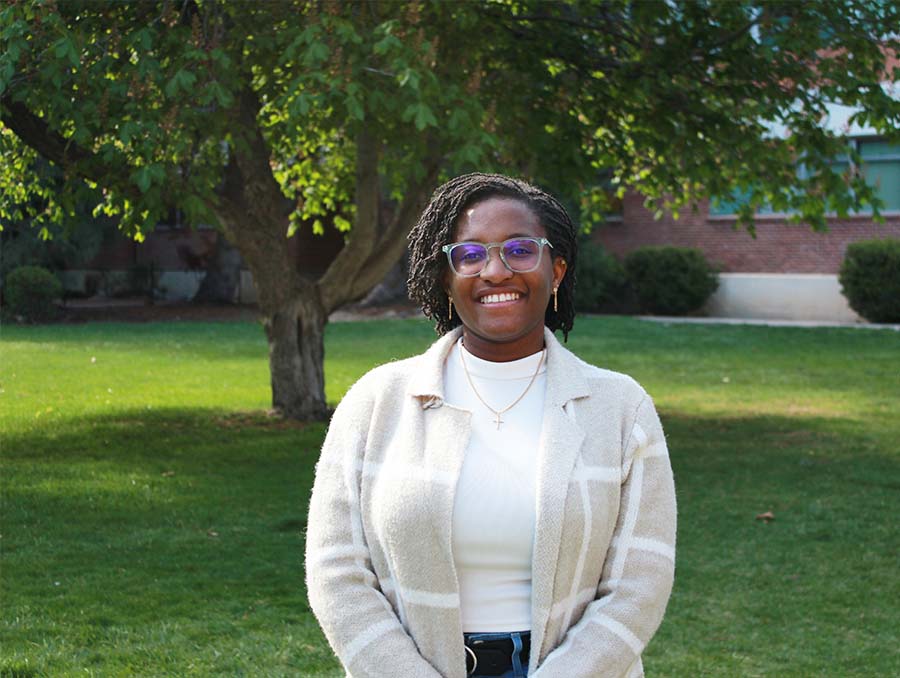 A smiling woman wearing glasses, a light plaid coat, and a white top stands outside on a green lawn with a large tree and brick building in the background.