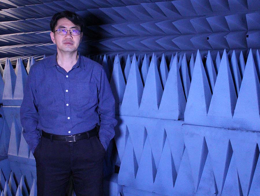 A man in a blue shirt stands in an anechoic chamber with blue, foam-covered wedge panels designed to absorb sound or electromagnetic waves.