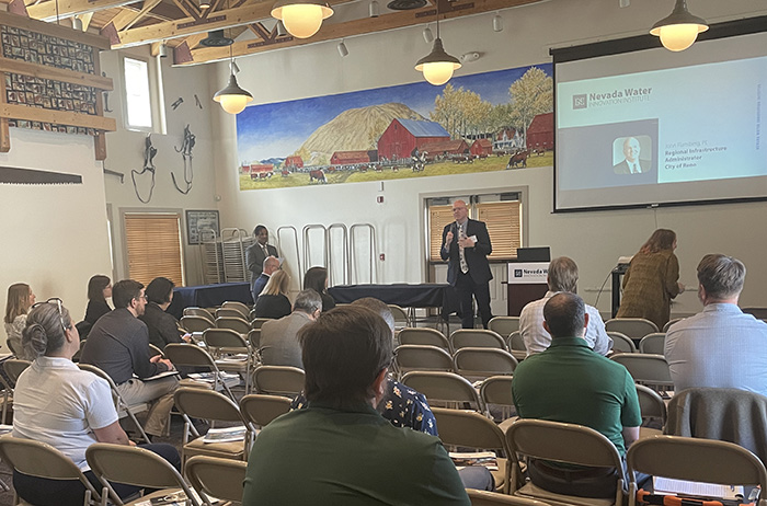 A presentation by Nevada Water at a conference with attendees seated facing a speaker at a podium, and a large mural of a rural landscape in the background.