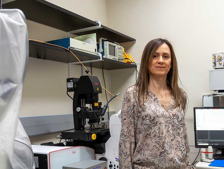 Woman in a tan print blouse stands in front of lab equipment, smiling at the camera.