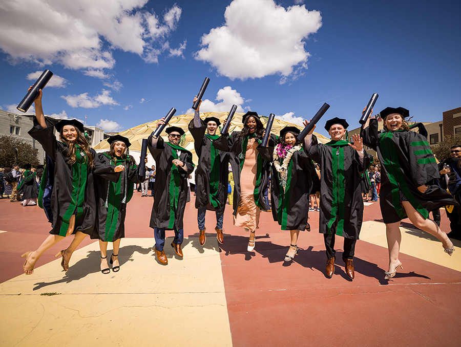 M.D. Class of 2025 graduates dressed in regalia outside the Pioneer Center jumping in the air with their diplomas in hand.