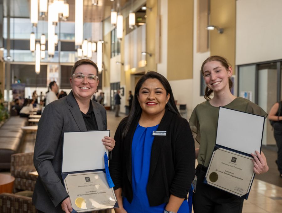 Three women smile for a photo. The women on the left and right hold certificates.