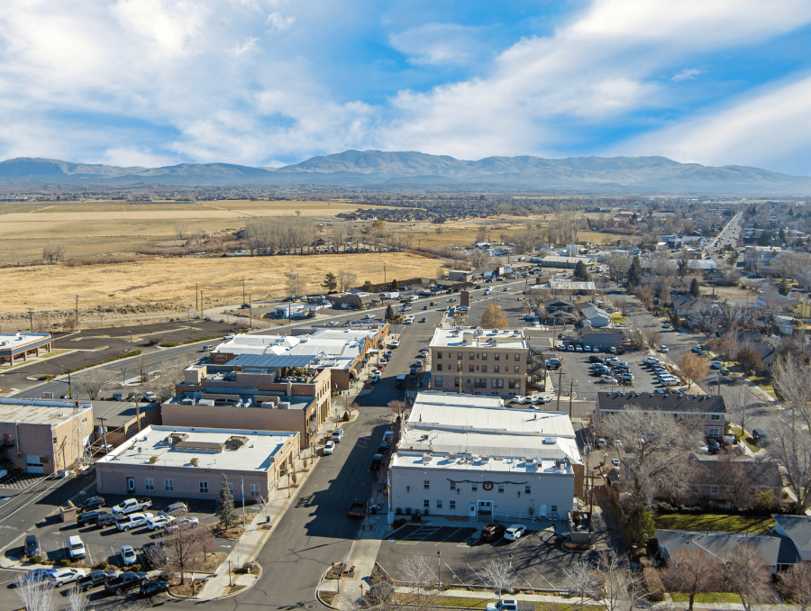Rows of buildings with cars parked out front, set against open grasslands under a bright blue sky with scattered white clouds.