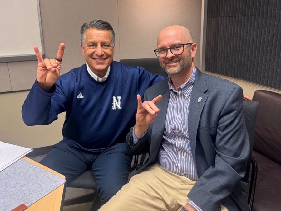 Brian Sandoval sitting next to Joe Nannini in the podcasting studio holding up Wolf Pack hand signs.