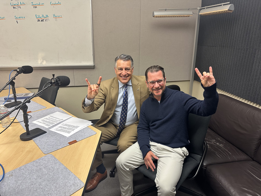 Brian Sandoval sitting next to Yani Dickens in the podcasting studio holding up Wolf Pack hand signs.