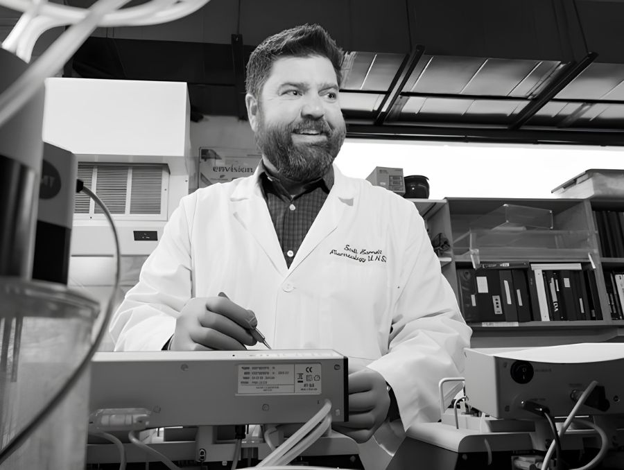 A black and white image of man in a white coat standing by an electronic machine. 