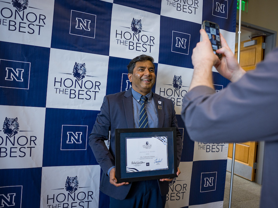 Professor Verma holding his certificate, smiling in front of an Honor the Best step and repeat backdrop while someone takes a cell phone photo. 