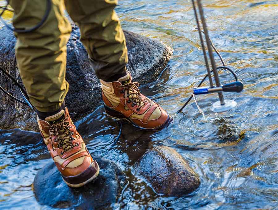 Person in hiking boots standing on rocks in a stream, using a metal detector to search the water.
