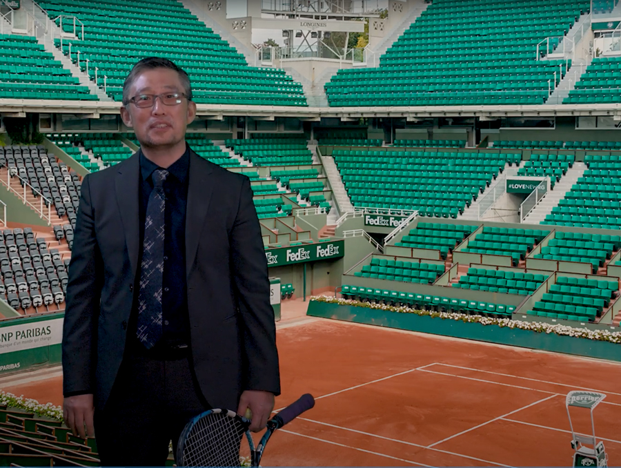 Dean Yun holding a tennis racket standing in front of a green screen image of the French Open court.