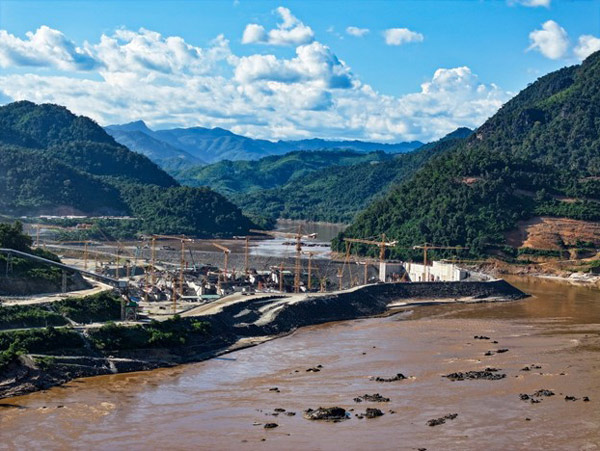 A dam under construction on a river that spans valleys between lush green hills.