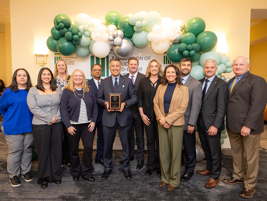 Group of individuals from UNR at the Association of Public and Land-grant Universities posing with an award in a room decorated with green and white balloons.
