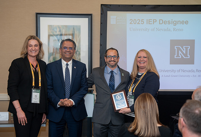 Four individuals standing at a ceremony, smiling as they are presented an award. A banner in the background reads "2025 IEP Designee, University of Nevada, Reno."