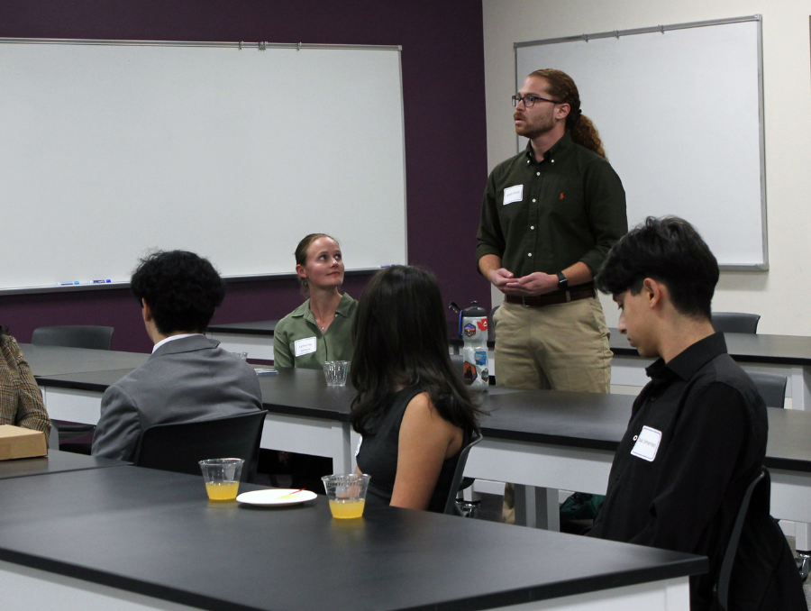 A student speaks to a small group during the Apple Mentorship Program reception in a classroom setting as attendees sit and listen attentively.